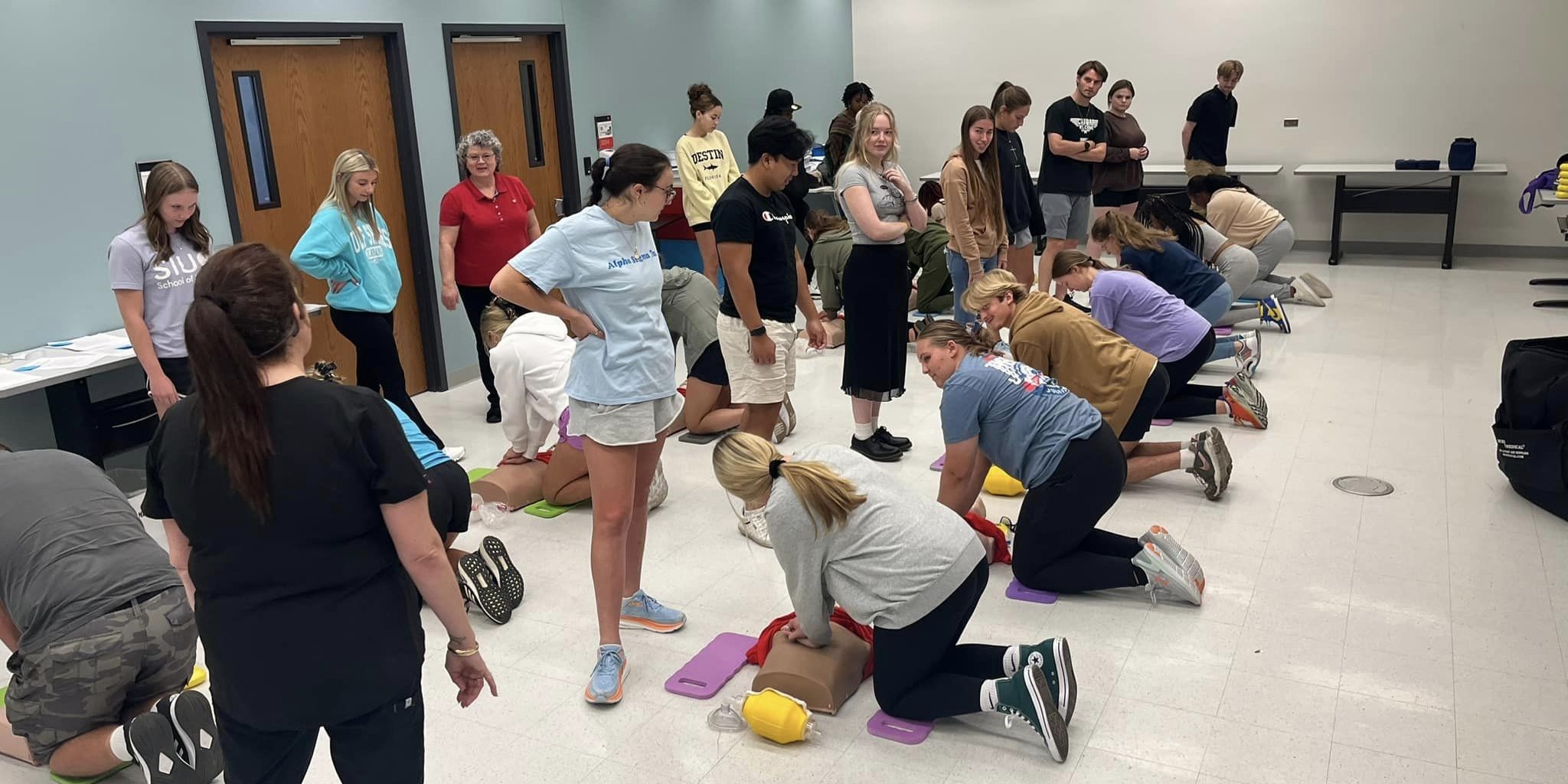 A group of college student learning how to perform CPR on training dummies while other students observe.
